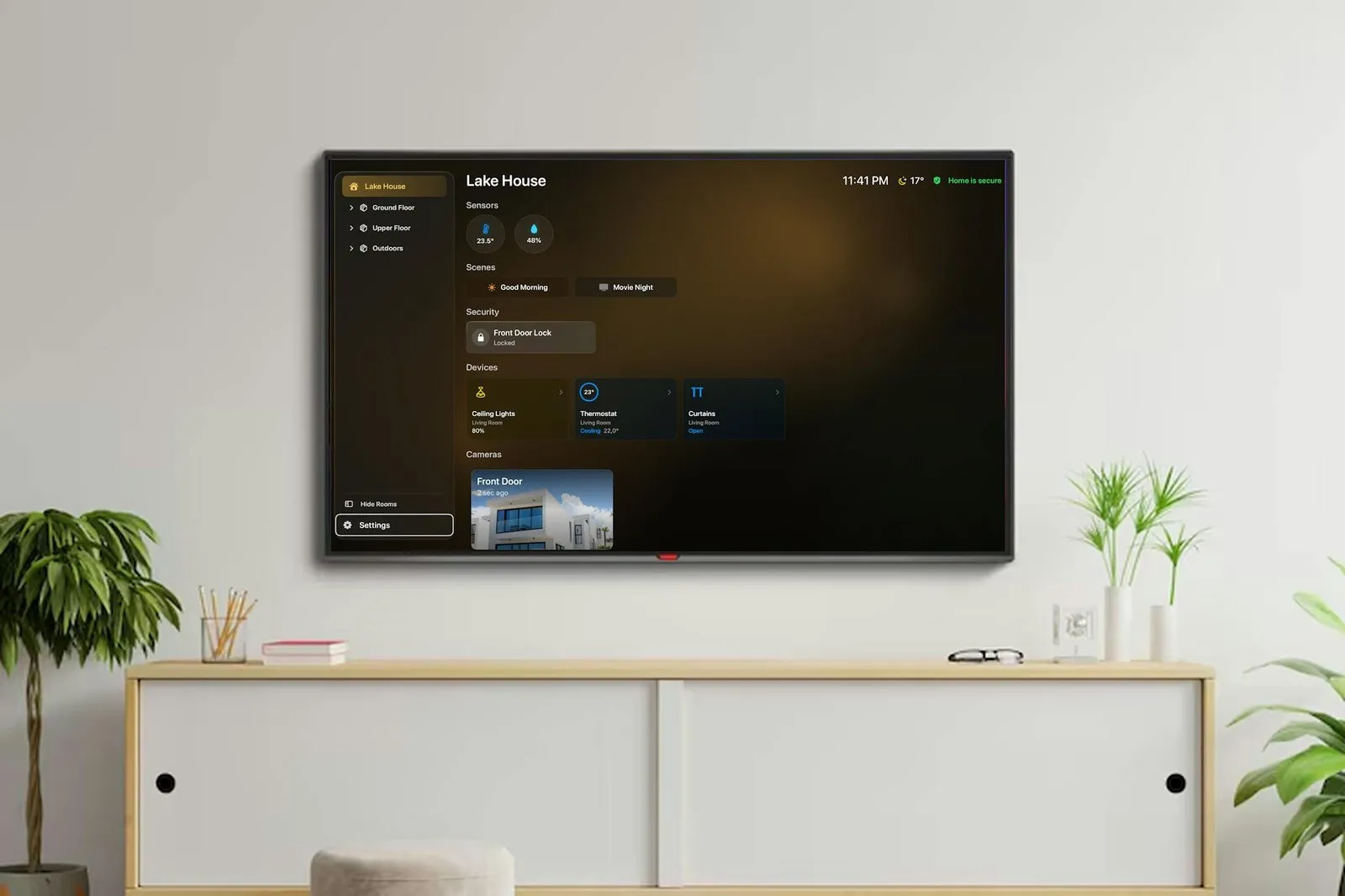 A wall-mounted TV showing the HomeDeck dashboard for a home called Lake House: a sidebar with Ground Floor, Upper Floor and Outdoors, sensor bubbles reading 23.5 degrees and 48 percent, Good Morning and Movie Night scenes, a locked Front Door Lock, Ceiling Lights at 80 percent, a Thermostat cooling to 22 degrees, open Curtains, and a Front Door camera tile, with the time, temperature and Home is secure in the top corner